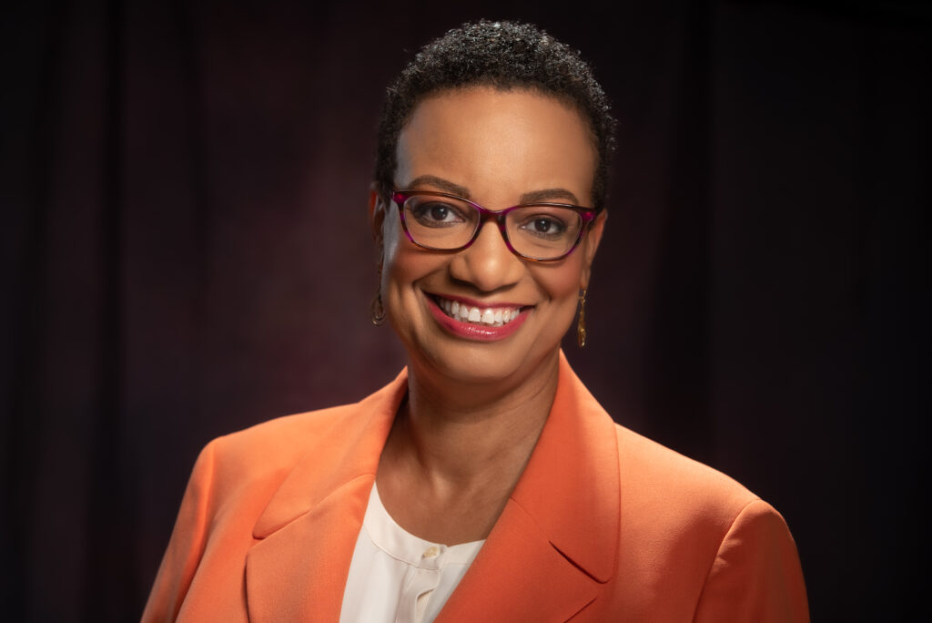 Headshot of Allison Demas, a woman of Afro-Caribbean decent, wearing an orange blazer with a white shirt, glasses, and earings.
