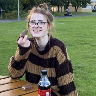 BRianna Ghey, sitting at a picnic table outside wearing a yellow and black stripped sweater smiling eating a piece of candy.